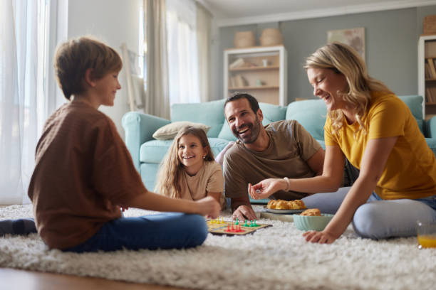 Family playing a board game together on the living room floor — parents, two children, all smiling and comfortable at home